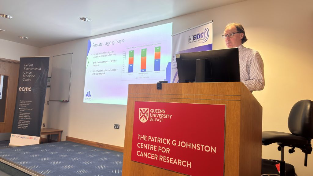 Damien Bennett from Northern Ireland Cancer Registry standing at the podium presenting data on cancer trials access in Northern Ireland 
